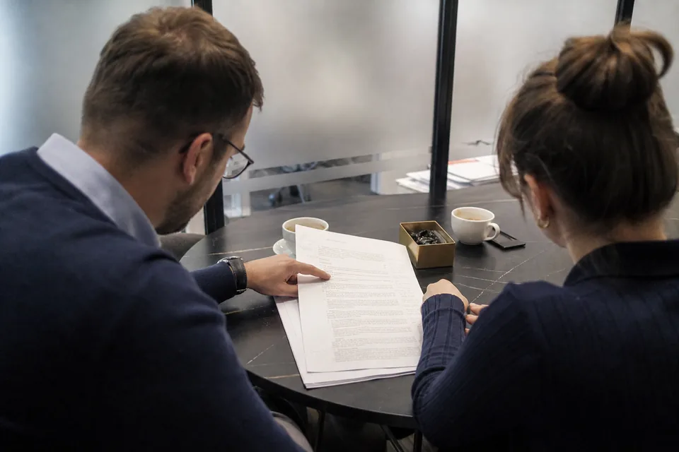 Dwie osoby przy stole w sali konferencyjnej czytają projekt umowy sprzedaży udziałów, obok leży wydruk sprawozdania finansowego